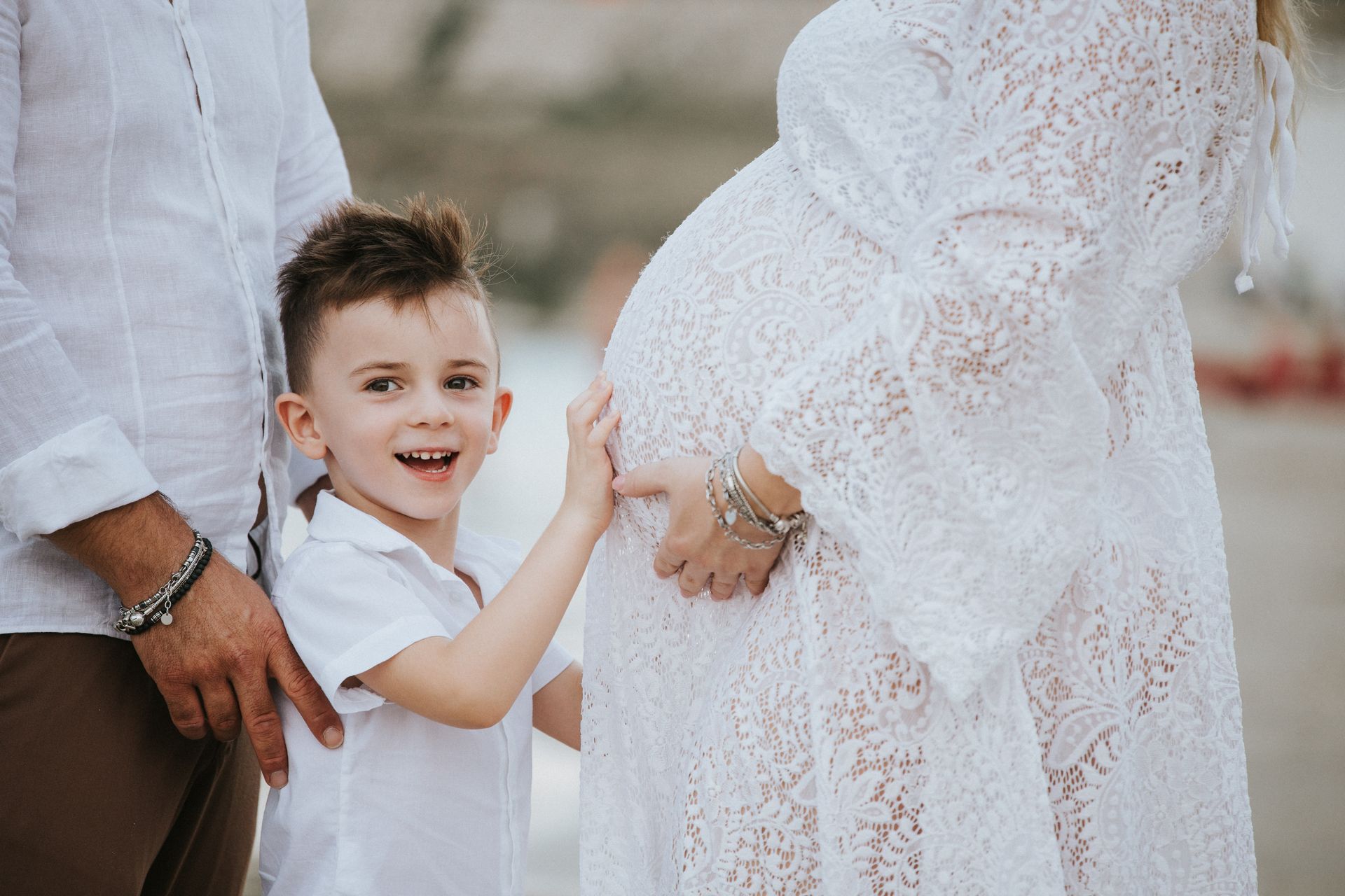 Bambino in camicia bianca sorride felice mentre appoggia le mani sul pancione della mamma