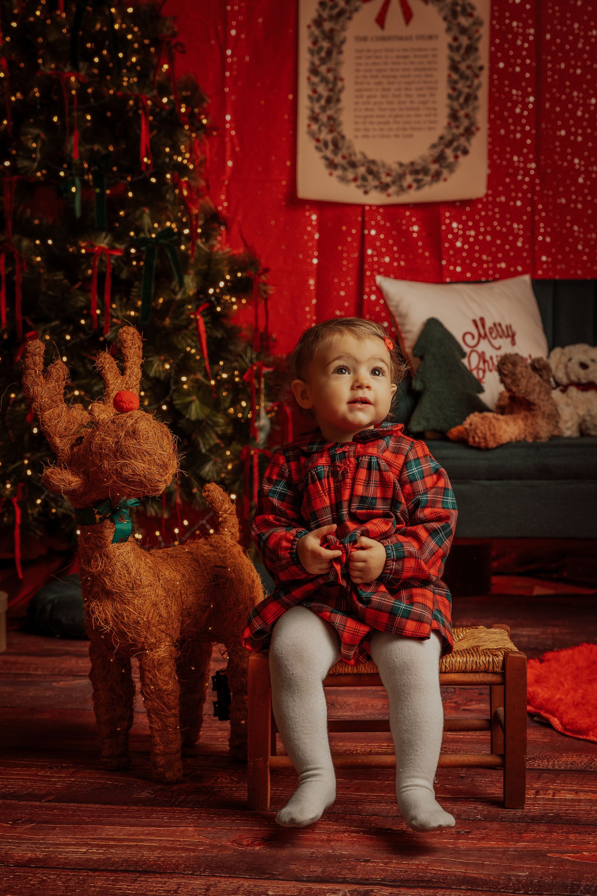 Bimba in abito scozzese seduta su una sedia di legno accanto alla renna di peluche, fra l'albero e le tende rosse