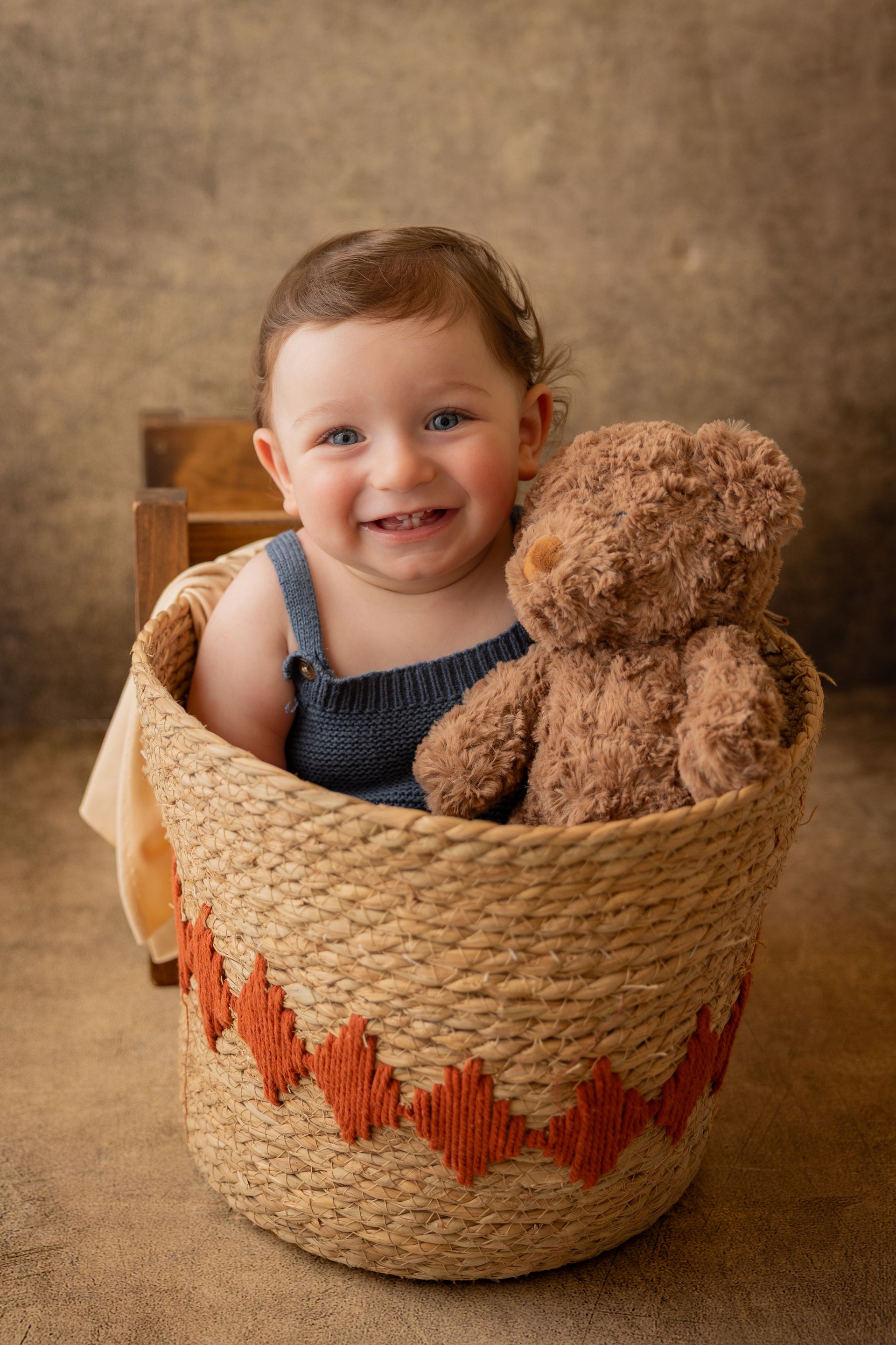 Bambino che ride a bocca aperta dentro un cesto di corda con cuori arancioni, accanto un orsetto morbido