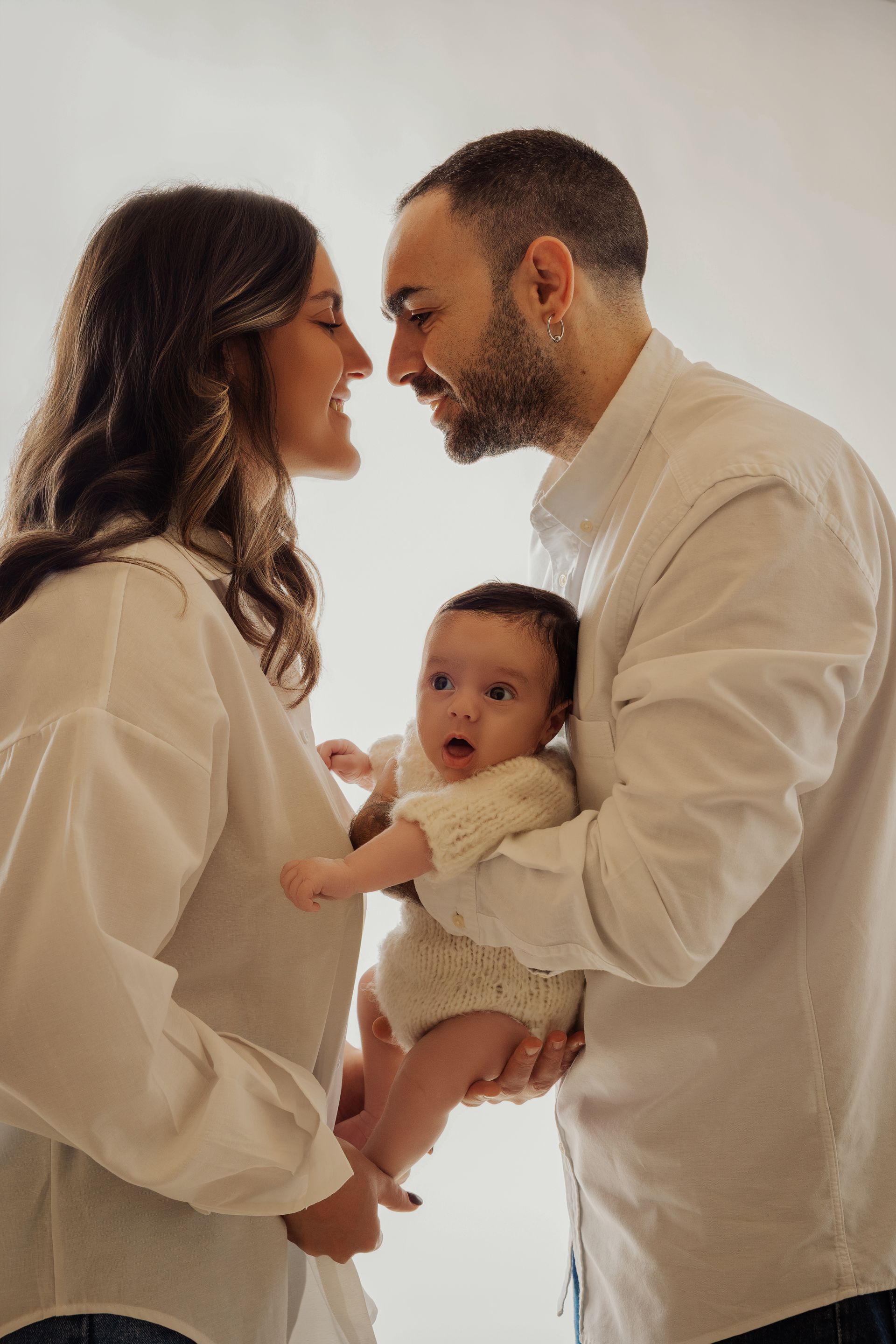 Coppia vestita di bianco si guarda fronte contro fronte tenendo la figlia neonata su fondo chiaro