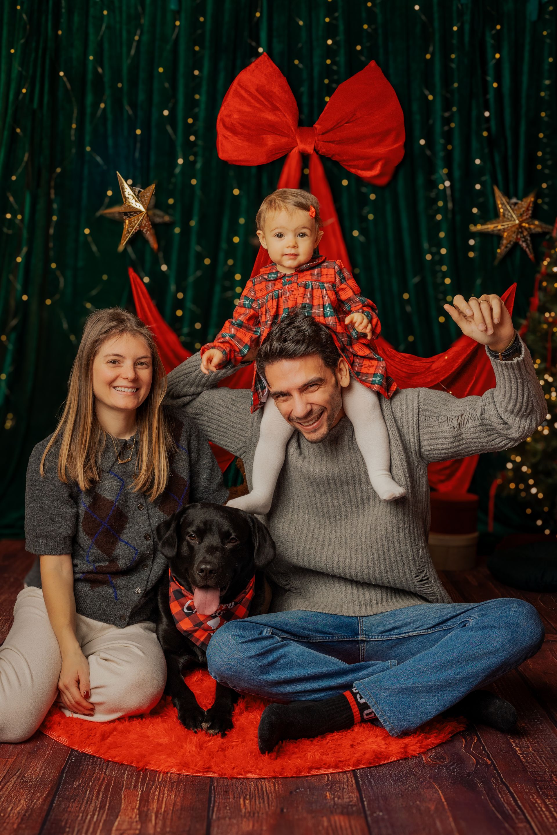 Mamma e papà con la bimba sulle spalle e il cane nero, davanti alle tende verdi e alle stelle del set natalizio