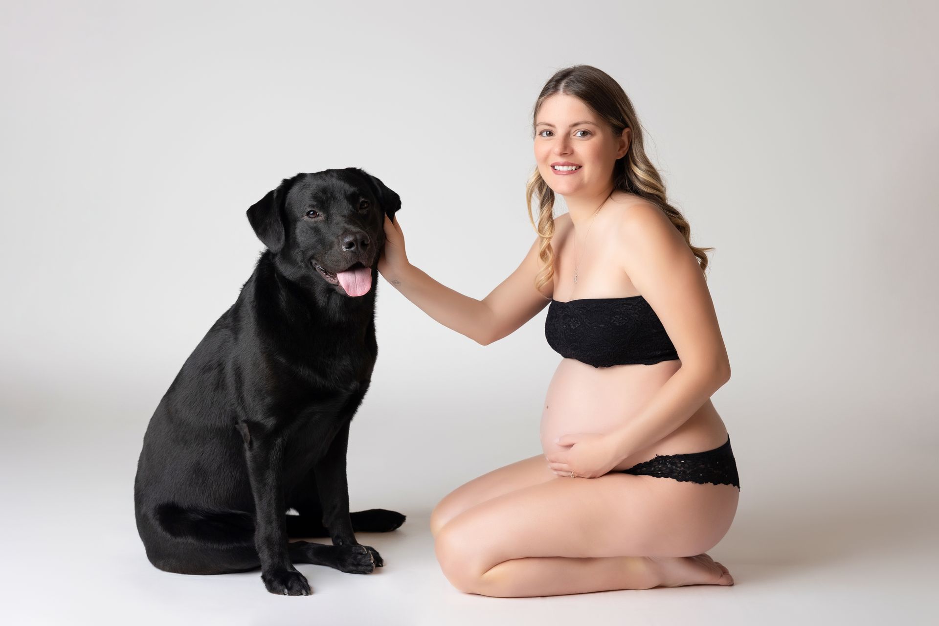 La futura mamma inginocchiata accarezza il labrador nero, su fondo bianco