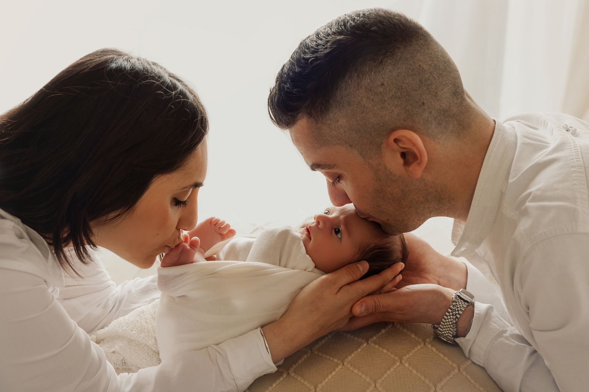 Papà bacia il neonato fasciato tenuto in braccio dalla mamma, entrambi vestiti di bianco