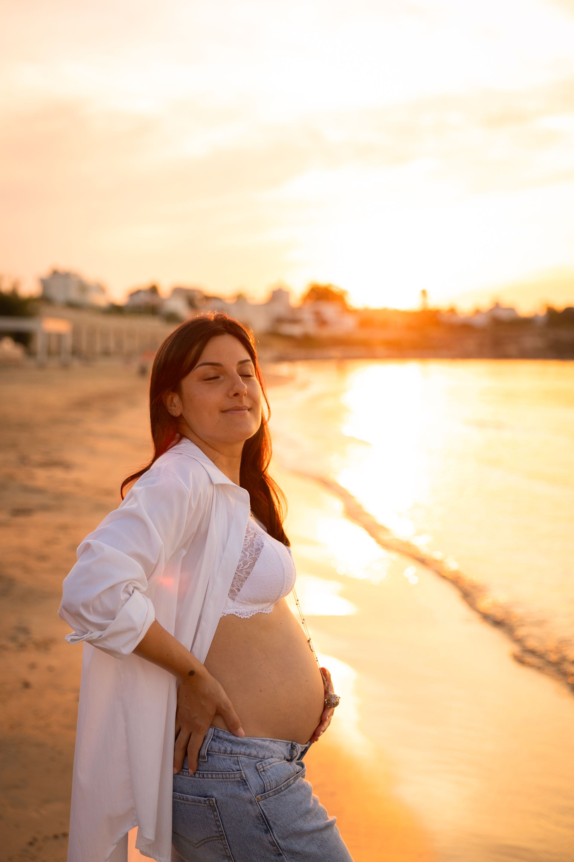 Futura mamma dai capelli ramati con camicia bianca aperta e jeans, sole basso sul mare
