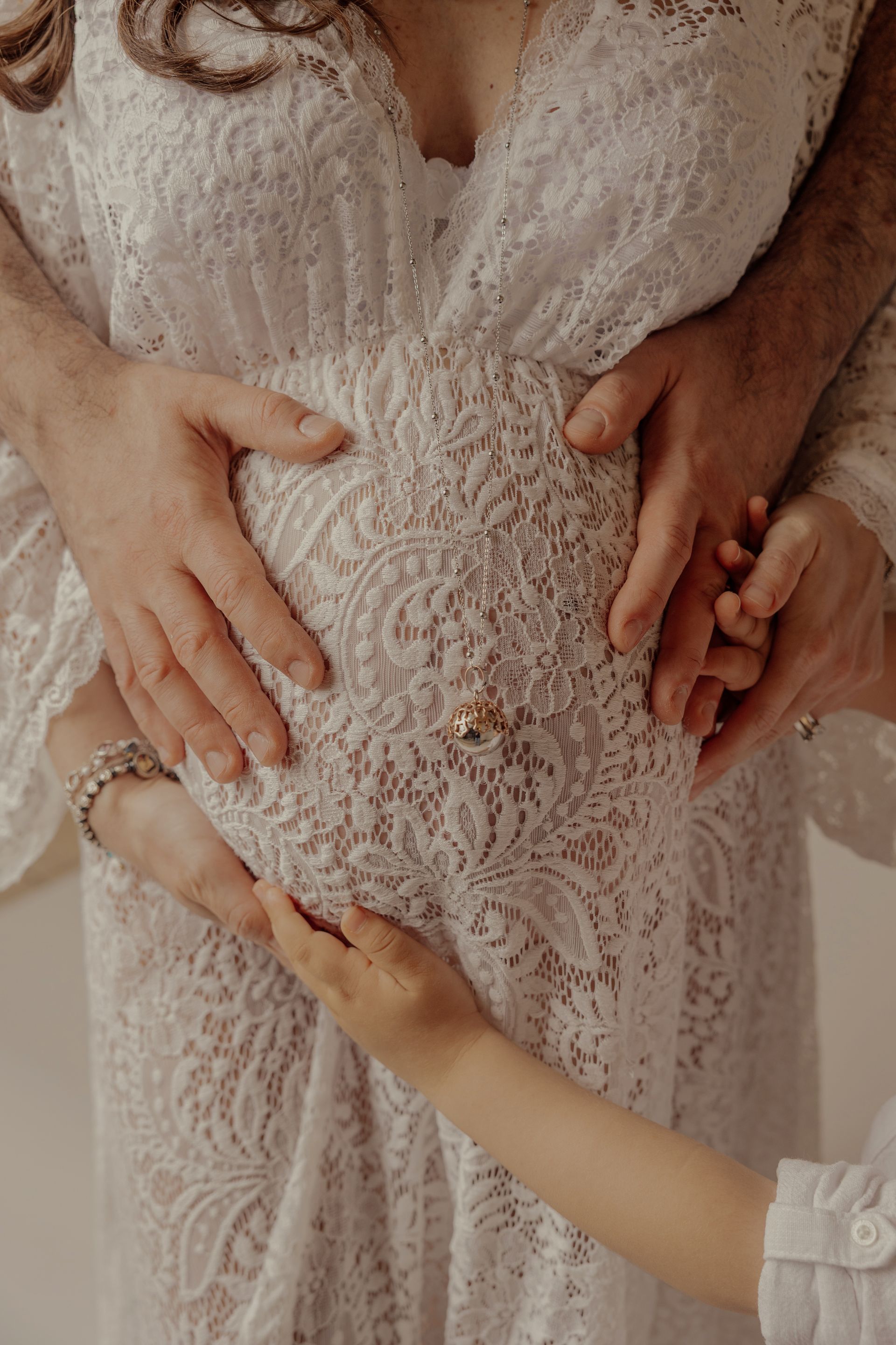 Le mani dei genitori e quella piccola di una bambina posate sul pancione vestito di pizzo