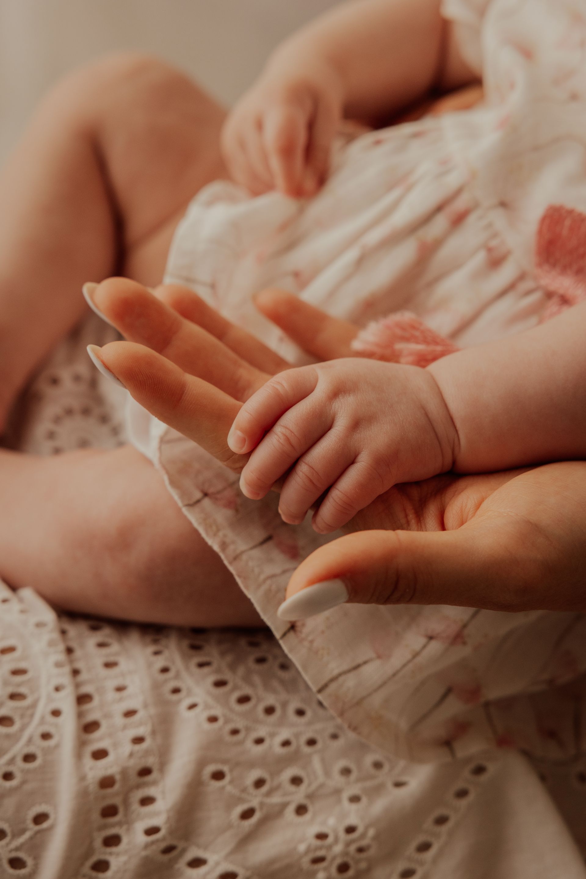 Manina di un bebè appoggiata sul palmo aperto della mamma, tessuti bianchi e rosa cipria intorno