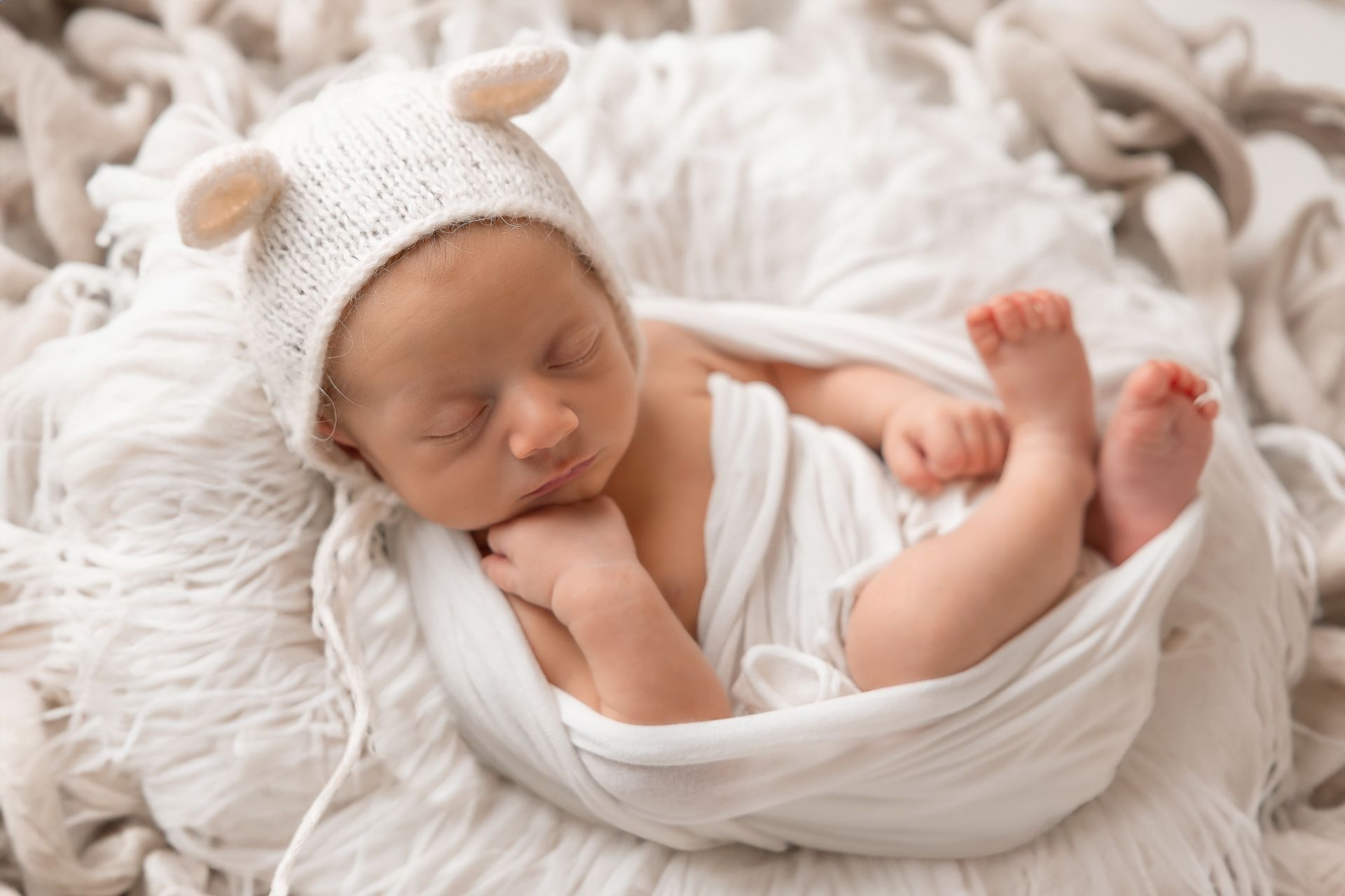 Neonato con cuffietta a orecchie dorme fasciato di bianco su un soffice tappeto crema