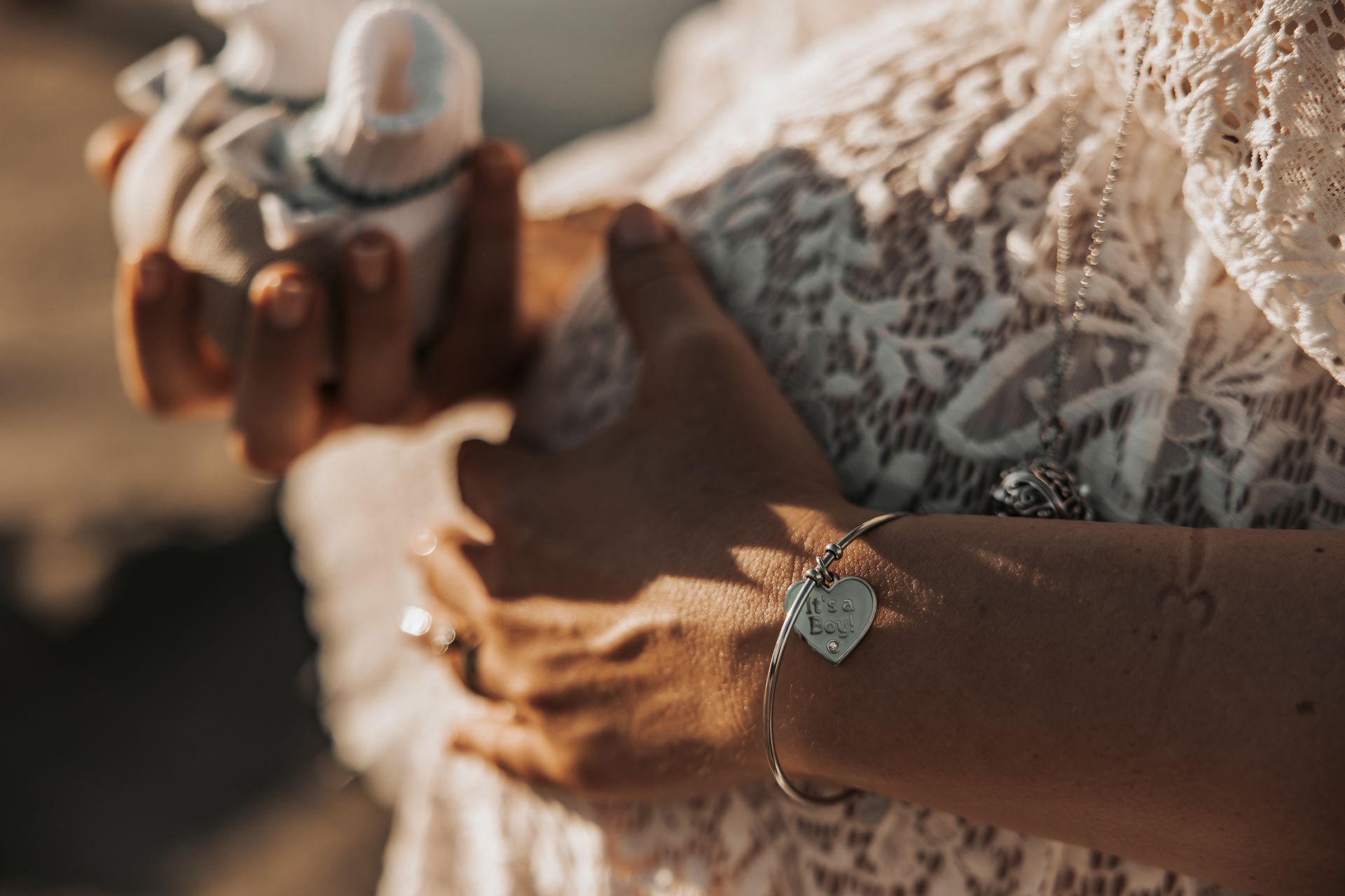 Mani che stringono minuscole scarpine sopra un abito di pizzo, bracciale con ciondolo a cuore